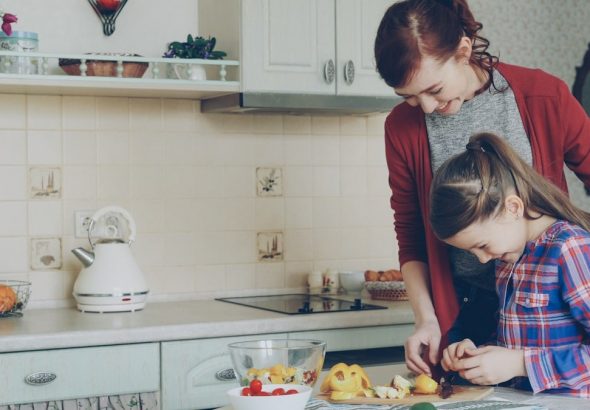 Young pretty mother teaching her cute daughter to cut vegetables properly. Little smiling girl cooking together with loving mom at home in modern kithcen